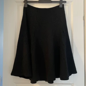 Svart blommig långkjol från Eiva Stils - Säljer en svart långkjol från Eiva Stils med snyggt blommigt strukturmönster. Kjolen har en utsvängd form och är tillverkad i en mix av modal, ull och akryl. Perfekt för dig som gillar en klassisk men ändå unik look.