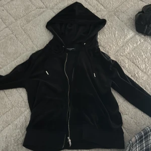 Svart hoodie från Juicy Couture - Svart hoodie från Juicy Couture i mjuk och glansig polyesterblandning. Tröjan har dragkedja framtill, huva med snörning och långa ärmar. Perfekt för en chill och trendig look.