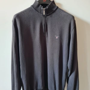 Mörkgrå half zip tröja från GANT strl L - Säljer en mörkgrå långärmad tröja från GANT i storlek L med ribbad hög krage och half zip-dragkedja. Tröjan har en liten broderad GANT-logga på bröstet och är tillverkad i 100% bomull.