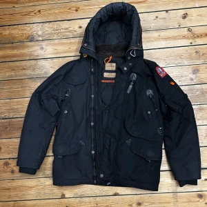 Svart Parajumpers vinterjacka M - Grym svart vinterjacka, en äkta Parajumpers Right Hand Masterpiece med huva, flera fickor och coola detaljer som patch på ärmen och gul rem bak. Jackan är vadderad och har dragkedja och knappar framtill. Perfekt för kalla dagar och riktigt snygg streetstil.