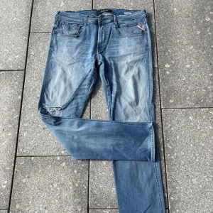Replay Anbass ljusblå jeans - Säljer ett par Replay Anbass jeans i ljusblå tvätt med slitningar och hål på ena knät. Klassisk femficksmodell med snygga detaljer och logga på fickan. Jeansen har normal passform och är tillverkade i mjukt bomullsjeansmaterial.