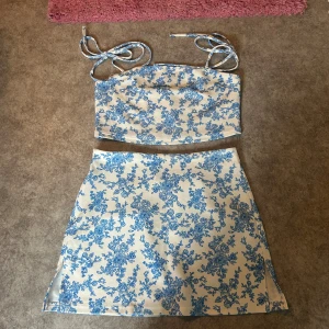 Vit-blå blommig kortkjol med dragkedja - Superfin kortkjol i vitt med blått blommönster. Kjolen har en lätt A-linjeform och stängs med dragkedja. Mönstret är riktigt somrigt och fräscht. Perfekt att matcha med crop tops eller linnen.