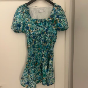 Blå blommig kortklänning från Zara - Supersöt kortklänning från Zara 🩵. Klänningen har puffiga korta ärmar, smockad midja och fyrkantig ringning. Storleken är S!