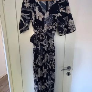 Mörkblå jumpsuit med blommigt mönster - Snygg jumpsuit i mörkblått med stora vita blommor och vackert knytband i midjan. Jumpsuiten har v-ringning, vida korta ärmar och ett flowy fall. Perfekt för dig som gillar en feminin och trendig stil.