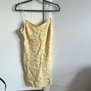 Gul blommig sommarklänning med smala band - Söt gul klänning med små blommor och tunna axelband. Den har en rak passform och är kort i modellen, perfekt för varma dagar. Klänningen är lätt och luftig, vilket gör den superbekväm att bära. Passar perfekt till sneakers eller sandaler.