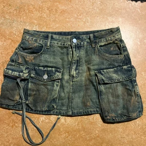Jeanskjol med stora fickor och snörning - Cool kort jeanskjol i mörkblå denim med slitna detaljer. Kjolen har stora cargofickor på sidorna, snörning och dragkedja på ena fickan samt klassiska jeansfickor fram och bak. Perfekt för en edgy streetstyle-look.