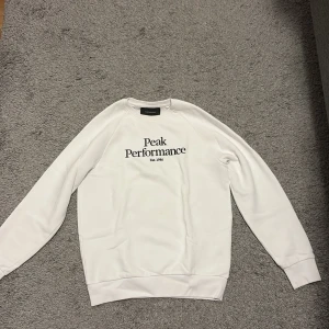 Vit sweatshirt från Peak Performance - Säljer en stilren vit sweatshirt från Peak Performance med svart logotyptryck på bröstet. 