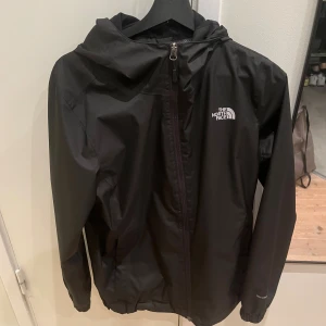 Svart vindjacka från The North Face - Svart vindjacka från The North Face med huva och dragkedja framtill. Jackan har en stilren design med logga på bröstet och ryggen, elastiska muddar och två sidofickor. Perfekt för blåsiga dagar och enkel att matcha med allt. Jackan är storlek S men passar även M om det skulle vara så!