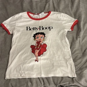 Betty Boop t-shirt med röd kant XS - Vit t-shirt från Clockhouse i storlek XS med röd kant vid hals och ärmslut. Framsidan har ett stort Betty Boop-tryck i svart och rött. T-shirten är kortärmad och har en klassisk, normal passform. Materialet är mjuk bomull, perfekt för dig som gillar retro och coola prints.