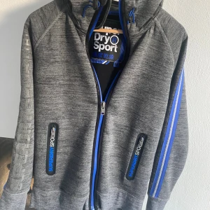 Grå hoodie Superdry Sport med blå detaljer - Snygg grå hoodie från Superdry Sport med blå dragkedja och blå ränder på axlarna. Tröjan har huva med snörning, tryck på ärmen och fickor med dragkedja. Perfekt för träning eller chill. 