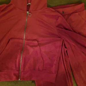 Rött mjukisset i velour  - Rött mjukisset i velour med dragkedja och stor huva. Setet har fickor och är tillverkad i ett mjukt, sammetsliknande material. Perfekt för dig som gillar en chill och avslappnad stil. Långärmad modell med normal passform.