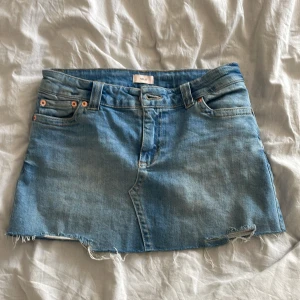 Blå jeanskjol från Nelly - Snygg blå jeanskjol från Nelly med råa kanter och klassiska fickor både fram och bak. Kjolen har bälteshällor och en enkel knappstängning framtill. Perfekt för dig som gillar en avslappnad och trendig stil.