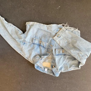 Ljusblå croppad jeansjacka - Säljer en ljusblå croppad jeansjacka med rå, fransig nederkant och två stora bröstfickor med knappar. Jackan har klassisk krage och långa ärmar. Perfekt för dig som gillar en avslappnad och trendig stil.