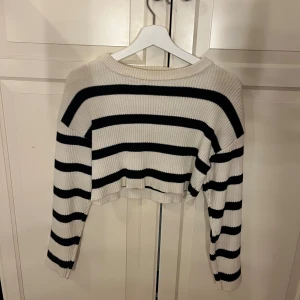 Randig croppad stickad tröja - Säljer en croppad stickad tröja med breda svarta och vita ränder. Tröjan har lång ärm, ribbad struktur och avslappnad passform. Perfekt för dig som gillar en chill och trendig stil.