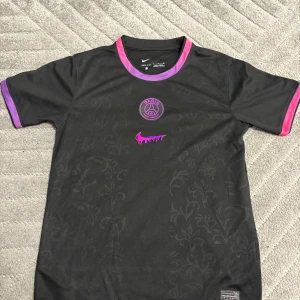 Svart PSG x Nike fotbollströja  - Snygg svart PSG fotbollströja från Nike med lila och rosa detaljer på krage och ärmslut. Tröjan har ett diskret blommigt mönster, Paris Saint-Germain-logga och en lila Nike swoosh med drip-effekt. Tillverkad i lätt Dri-FIT material som andas.