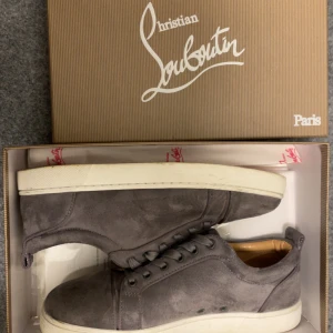 Grå mockasneaker Christian Louboutin - Snygga grå sneakers från Christian Louboutin i mjuk mocka med klassisk röd sula. Skorna har rund tå, snörning och en vit platt sula. Insidan är fodrad i beige läder för extra komfort. Perfekta för dig som vill ha en stilren och lyxig look.