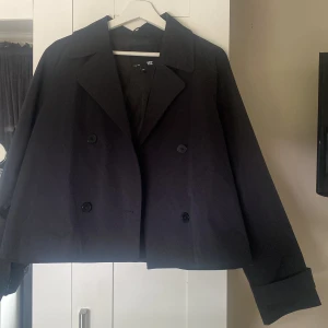 Svart croppad rock från 157, stl M - Snygg svart croppad rock från 157 med dubbelknäppning och bred krage. Jackan har långa ärmar med knappar vid ärmslut och en stilren, minimalistisk look. Perfekt för dig som gillar klassiska plagg med modern twist.