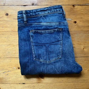 Blå jeans från Tiger of Sweden - Snygga blå jeans från Tiger of Sweden med klassisk femficksdesign och diskreta slitningar. Jeansen har raka ben och normal passform, tillverkade i bomull. Vid snabb affär kan priset ändras