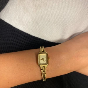 Guldfärgad rektangulär klocka BOVOU - Stilren guldfärgad klocka från BOVOU med rektangulärt ur och länkarmband i metall. Urtavlan är vit med minimalistiska svarta markeringar och två. Går att justera storlek hos urmakare. Säljs på hemsidan för 1395 kr.