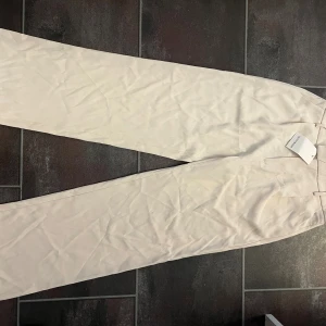 Beiga kostymbyxor från Outfitbook - Stilrena beiga kostymbyxor från Outfitbook i rak modell. Byxorna har hög midja, bälteshällor och diskreta pressveck framtill. Perfekta för en clean och trendig look. Materialet känns mjukt och har en lätt glans. Helt oanvända! 