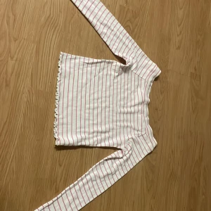 Randig långärmad tröja från Kappahl - Vit långärmad tröja från Kappahl med tunna rosa ränder. Tröjan är ribbad och har en söt volangkant nertill. Perfekt för dig som gillar en enkel men ändå söt stil.