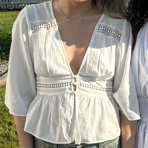 Vit blus med spets och knyt - Säljer en luftig vit blus med v-ringning, dekorativa spetsdetaljer och knyt framtill. Blusen har trekvartsärmar och ett lätt rynkat fall nedtill. Perfekt för varma dagar och ger en romantisk vibe.