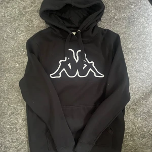 hoodie från Kappa - Svart hoodie från Kappa i storlek small med stor vit Kappa-logga på bröstet. Tröjan har huva med snörning och en klassisk känguruficka framtill. Perfekt för en avslappnad och sportig stil.