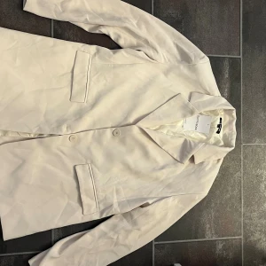 Beige kavaj från Outfittery - Snygg beige kavaj från Outfittery med klassisk krage, två knappar framtill och fickor med lock. Ärmslutet har fyra dekorativa knappar och insidan är helfodrad i blankt tyg. Perfekt för en clean och stilren look. Helt oanvänd 