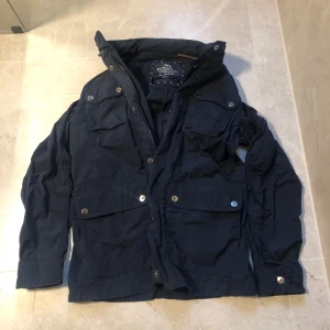 Mörkblå filed jacket från bondelid - Snygg mörkblå vindjacka från Bondelid med flera praktiska fickor framtill och hög krage. Jackan har både dragkedja och knappar, vilket ger en cool look och extra skydd mot vinden. Perfekt för dig som gillar stilrena och funktionella plagg.