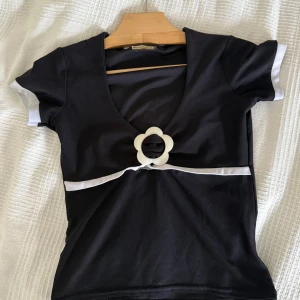 Svart topp med blomdetalj Nova Fashion - Supersöt  vintage  topp från Nova Fashion med korta ärmar och vit kant längs ärmslut och under bysten. Djup v-ringning och en stor vit blomformad detalj framtill som ger en unik retrovibe. Mycket bra skick  uppskattad storlek S