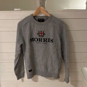 Grå stickad tröja från Morris - Snygg grå stickad tröja från Morris Gents med broderad logga och brittisk flagga på bröstet. Tröjan har rund halsringning, ribbade muddar och är långärmad. Perfekt för dig som gillar klassisk och stilren look.