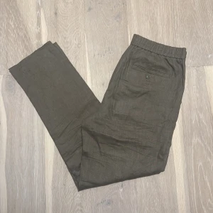 Olivgröna linnebyxor från These Glory Days - Snygga olivgröna linnebyxor från These Glory Days i storlek 48(M). Byxorna har elastisk midja bak, raka ben och diskreta bakfickor med knapp. Perfekta för en chill och stilren look under varmare dagar. Knappt använda och i väldigt bra skick.