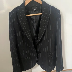 Svart randig kavaj från H&M - Svart kavaj från H&M med tunna vita ränder i klassiskt snitt. Kavajen har två knappar framtill, fickor med lock och helfodrad insida. Perfekt för en stilren och trendig look. Materialet är mjukt och följsamt, vilket gör den bekväm att bära hela dagen.