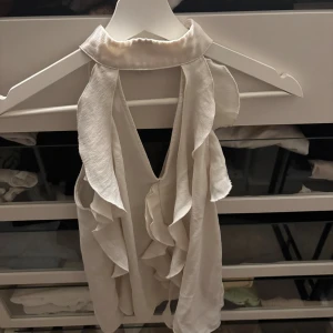 Vit volangblus med öppen rygg - Supersöt vit blus med volangdetaljer och hög krage. Toppen har en djup v-ringning framtill och öppen rygg, vilket ger en riktigt snygg och trendig look. Perfekt för dig som gillar romantiska vibes och flowiga material.