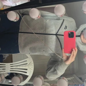 Grå Nike Tech hoodie med dragkedja - Säljer en grå hoodie från Nike med svart logga på bröstet och hel dragkedja framtill. Tröjan har huva, långa ärmar och svarta detaljer längs sömmarna. Perfekt för chill dagar eller när du vill ha en sportig look. Skit snygg, säljer ENDAST för den inte kommer till användning längre 🥰 Använt 3 gånger ‼️☺️