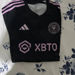 Inter Miami Messi #10 fotbollströja - Svart kortärmad Inter Miami fotbollströja från Adidas med rosa detaljer och tryck. Messi #10 på ryggen, klubbmärke på bröstet och sponsorlogga framtill. Tillverkad i lätt och ventilerande polyester, perfekt för match eller träning.