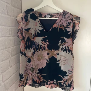 Blus - Bommig blus från Saint Tropez i storlek S. Blusen har v-ringning och volangiga ärmlösa axlar. Under blomtyget är det ett mörkare tyg som man ser på sista bilden.  Använd någon enstaka gång men som ny. 