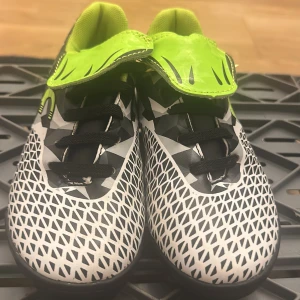 Svarta och vita fotbollsskor med neon - Säljer ett par coola fotbollsskor med svart och vitt grafiskt mönster och detaljer i neonlimegrönt. Skorna har snörning, platt sula och en bred plös i syntetmaterial. Insidan är fodrad i matchande limegrönt tyg för extra komfort. Perfekta för dig som vill sticka ut på planen!