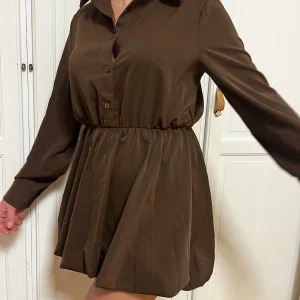 Brun skjortklänning från Dolce Vita - Kort brun skjortklänning från Dolce Vita med lång ärm, krage och knappar framtill. bubblig nedre del Klänningen har markerad midja med resår och ett lätt glansigt tyg som ger en snygg look. Perfekt för dig som gillar stilrena och trendiga plagg.