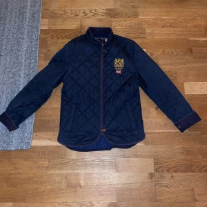 Morris quiltad jacka marinblå  - Quiltad jacka från Morris i marinblått med guldbroderad logga och brittisk flagga på bröstet. Jackan har ståkrage med manchesterdetaljer, två fickor fram och stängs med dragkedja. Snyggt rutmönster över hela jackan och muddar med knapp vid ärmslut. Perfekt för dig som vill ha en stilren jacka till vardags. Storlek xs. Skick 7/10
