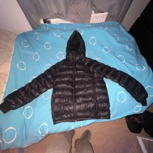 Svart dunjacka Peak Performance M - Svart dunjacka från Peak Performance i storlek M. Jackan har huva, ribbade muddar och dragkedja framtill. Den är quiltad och har en snygg, sportig look som passar perfekt när det är kallt ute. Materialet är syntet och dun, vilket gör den både lätt och varm.