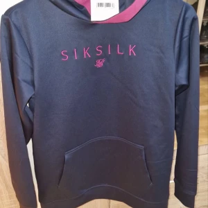 Mörkblå hoodie från SikSilk - Snygg mörkblå hoodie från SikSilk med rosa broderad logga på bröstet. Tröjan har huva med rosa insida och en stor magficka framtill. Perfekt för dig som gillar streetwear och vill ha en stilren look.