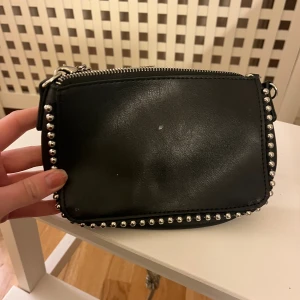 Svart axelväska med kedja och nitar - Snygg svart axelväska i skinnimitation med silverfärgade nitar längs kanterna och en kedja som axelrem. Väskan har dragkedja upptill och en stilren, rektangulär form som passar perfekt för att bära det viktigaste. Perfekt accessoar för att lyfta din outfit.