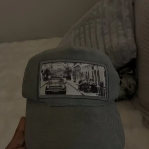 Grå meshkeps med vintagefoto - Säljer en grå keps med mesh på sidorna och baksidan. Framtill finns ett stort svartvitt vintagefoto med bil och stadsmiljö som ger kepsen en unik vibe. Perfekt för dig som gillar streetstyle och vill sticka ut med coola detaljer.