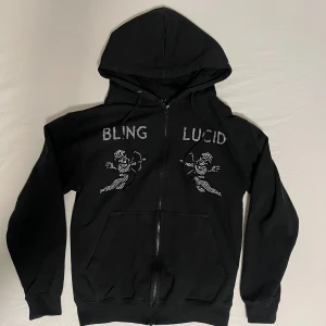 Svart rhinestone hoodie från Bling Lucid - Svart rhinestone hoodie från BlingLucid med strassdetaljer och motiv av två änglar på bröstet. Tröjan har huva med snörning, dragkedja framtill och stora fickor. Texten BLING LUCID i strass ger en extra blingad vibe. Perfekt för dig som gillar streetstyle.