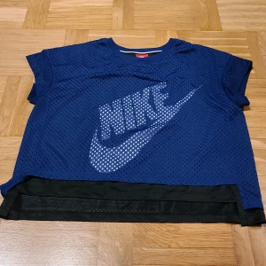 Blå croppad mesh-topp från Nike - Säljer en blå croppad mesh-topp från Nike med stort vitt logotryck framtill och 'JUST DO IT.' på ryggen. Toppen har korta ärmar och svart innerlager, perfekt för träning eller sportiga outfits. Luftigt material i polyester för extra komfort.