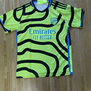 Arsenal kortärmad fotbollströja gul - Säljer en Arsenal fotbollströja från Adidas i neon-gul med svarta och blå detaljer. Tröjan har ett unikt randigt och prickigt mönster, korta ärmar och klubbmärke på bröstet. Materialet är lätt och ventilerande polyester, perfekt för träning eller match.