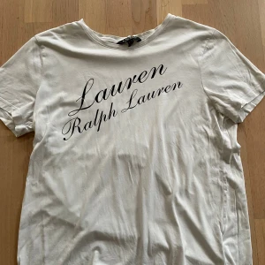 Vit t-shirt från Ralph Lauren - Vit t-shirt från Ralph Lauren med svart logotyptryck över bröstet. Klassisk rund halsringning och korta ärmar. Tillverkad i mjuk bomull för en skön och avslappnad känsla. Perfekt till jeans eller shorts för en clean och stilren look. Aldrig andvänt