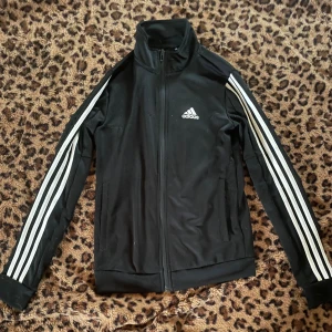 Svart Adidas track jacket 2XS - Svart Adidas track jacket med klassiska vita ränder längs ärmarna och dragkedja framtill. Jackan har hög krage, två fickor och Adidas-logga på bröstet. Perfekt för en sportig och avslappnad stil.