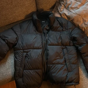 Svart pufferjacka från Zara - Säljer en svart pufferjacka från Zara med hög krage och dragkedja framtill. Jackan har quiltad design och en patch på ena ärmen med texten 'dnwr'. Perfekt för kalla dagar och har en clean, modern look.